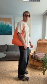 Saco tote laranja e texturizado com alças de ombro, usado sobre uma camiseta bege e jeans escuros. Apresenta um design casual adequado para o uso diário.