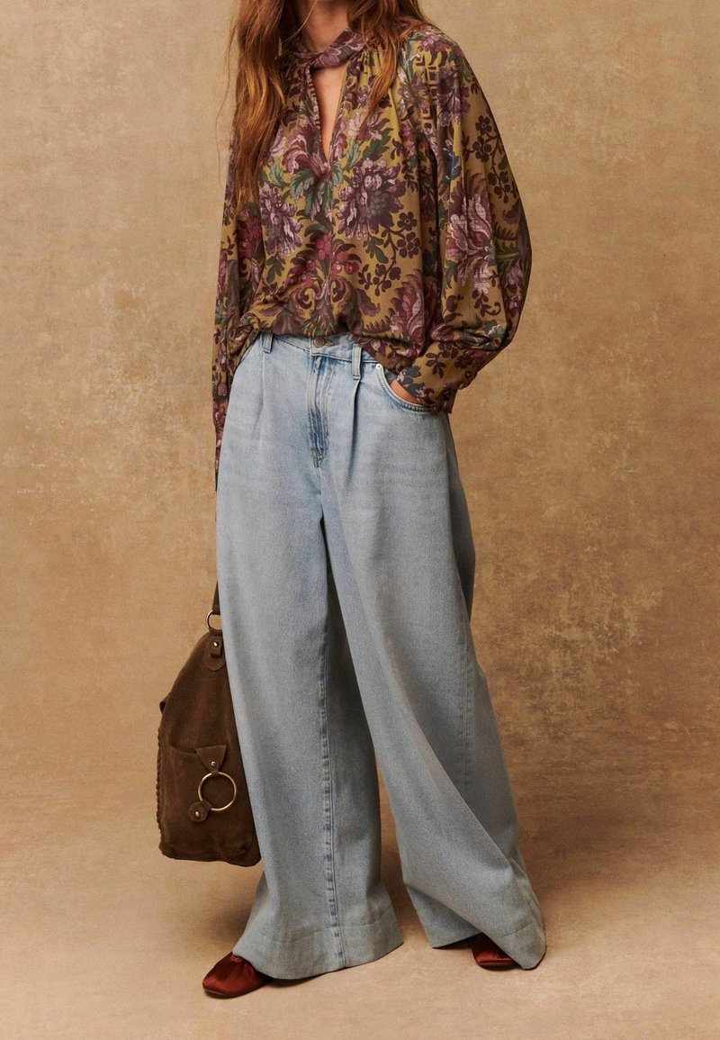 Personne portant un jean bleu clair ample, un chemisier à manches longues à motifs floraux dans des tons marron et violet, tenant un sac marron, debout sur un fond beige.