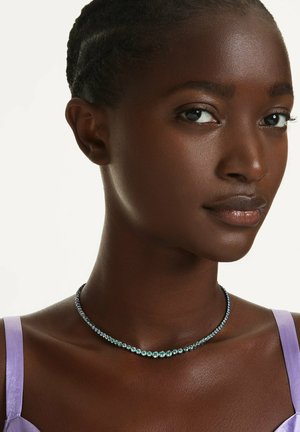 Primo piano di donna che indossa una maglia viola e una collana con gemma azzurra-verde, rivolta leggermente a destra con espressione neutra.