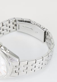Montre en acier inoxydable argenté avec un bracelet texturé et un fermoir argenté. Le cadran rond présente un fond blanc et trois sous-cadrans, embellis de pierres.