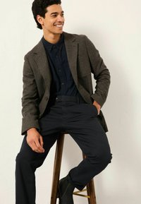 Braun gefärbter Hahnentrittmuster-Blazer über einem navyblauen Hemd, kombiniert mit dunklen Hosen und Stiefeletten. Mann sitzt auf einem Holzschemel vor einem einfarbigen Hintergrund.