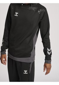 Haut de survêtement noir avec texture à pois sur les épaules, accents gris sur les manches et sur les côtés, arborant un logo sur la poitrine et sur le pantalon.