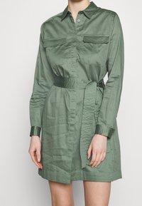 Donna che indossa un vestito camicia corto con maniche lunghe, di colore verde oliva, dotato di cintura, tasche sul petto e polsini con bottoni, su uno sfondo semplice.