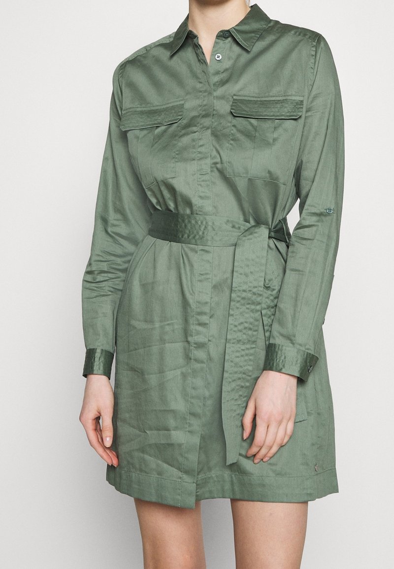 Donna che indossa un vestito camicia corto con maniche lunghe, di colore verde oliva, dotato di cintura, tasche sul petto e polsini con bottoni, su uno sfondo semplice.