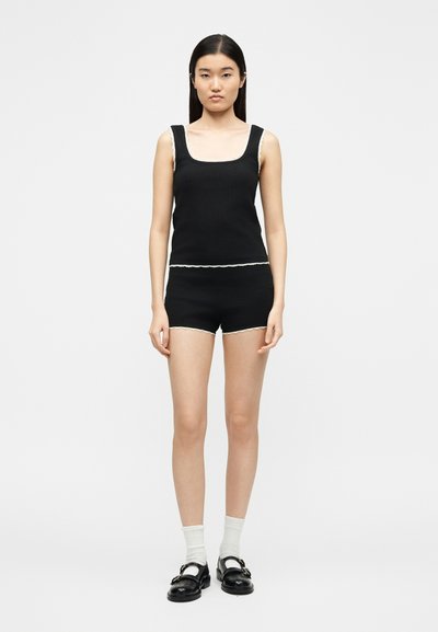 Jeune femme portant un haut noir sans manches et un short assorti avec un liseré blanc, associés à des chaussettes blanches et des mocassins noirs, debout sur un fond uni.