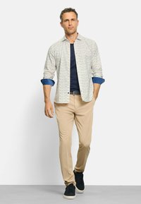 Hemd mit knöpfbarem Kragen in weißer Basis und buntem abstraktem Muster, kombiniert mit einem marineblauen T-Shirt und beigen Hosen, abgerundet mit schwarzen Sneakers.