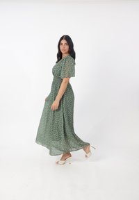 Grünes Blumenkleid im Maxistil mit kurzen Ärmeln, elastischem Taillenbund, fließender Textur und goldenen Blumenmustern. Kombiniert mit beigen High-Heel-Sandalen.