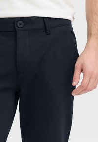 Pantaloni blu navy con una texture liscia, dotati di chiusura con bottone e design a vita piatta. Una mano è posizionata accanto alla cintura.