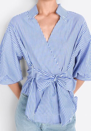 Niebiesko-biała, pionowo prążkowana bluzka na zakładkę z szerokimi rękawami i dużą kokardą z przodu, zestawiona z jasnoniebieskimi dżinsami.