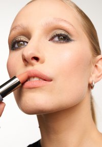 Nude szminka w srebrno-szarym opakowaniu nałożona na usta; modelka ma neutralny makijaż z złotym cieniowaniem i prostą obrączką na uchu.