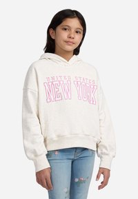 Ragazza con capelli scuri che indossa un hoodie crema con la scritta "Stati Uniti New York" in rosa e jeans chiari con ricami floreali.