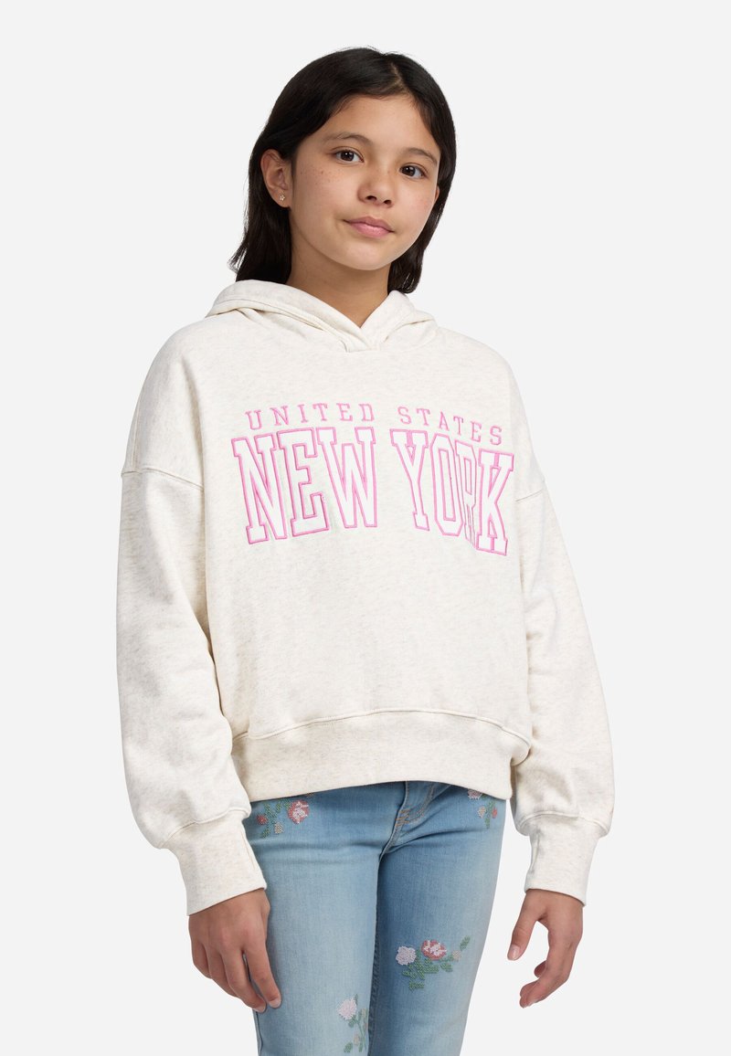 Ragazza con capelli scuri che indossa un hoodie crema con la scritta "Stati Uniti New York" in rosa e jeans chiari con ricami floreali.