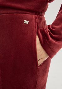 Vêtement en velours bourgogne avec une poche latérale et un accent logo argenté près de la taille, en tissu doux et texturé.