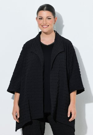 Uśmiechnięta kobieta z zaczesanymi do tyłu włosami nosi długą, fakturowaną czarną kurtkę i pasujące spodnie na jednolitym jasnym tle.