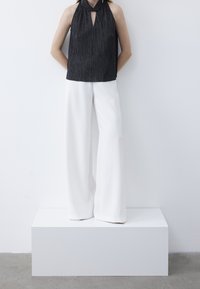 Osoba oblečená v bezrukávém černém halter topu a širokých bílých kalhotách stojí na bílé podložce před bílou zdí.