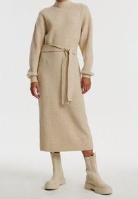 Robe midi en tricot côtelé beige avec des manches longues, un col montant et une taille à nouer. Associée à des bottines à plateforme crème.