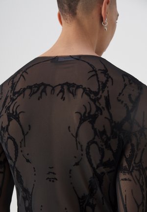 Rückansicht einer Person, die ein durchsichtiges schwarzes Oberteil mit kunstvollen samtartigen abstrakten Blumenmustern trägt.