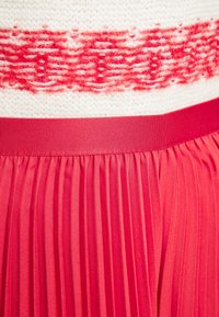 Punamainen laskostettu hame, jossa on sileä vyötärö, ja valkoisen neulotun paidan, jossa on punavalkoinen kuviolaita. Kangas on pehmeä ja kevyt.