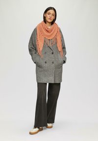 Manteau en laine gris à boutons pour double boutonnage, associé à une écharpe orange à franges. Un pantalon foncé évasé et des baskets beiges complètent la tenue.