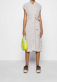 Nainen, joka käyttää vaaleaa kukallista nappiomistakin mekkoa, jossa on vyötärösolki, pitää käsissään kirkkaan keltaista käsilaukkua ja käyttää mustia platform-sandaaleja.