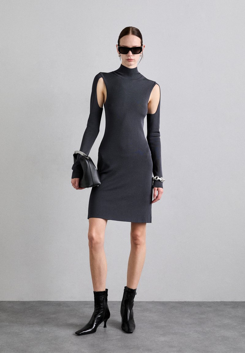 Grijze, nauwsluitende jurk met lange uitsnijdingen aan de mouwen en een hoge hals. Gepaarde met zwarte, puntige enkellaarsjes en een zwarte handtas.