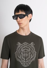 Gafas de sol rectangulares negras con lentes oscuros, llevadas con una camiseta verde oscuro que presenta un gráfico detallado de la cabeza de un león en gris.