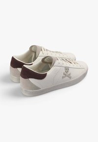 Zapatillas blancas de cuero con un acento de ante beige y cuero marrón en el talón. Presentan una suela de goma texturizada y un diseño de calavera bordado.