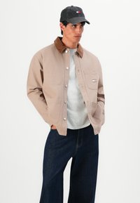 Man wearing beige jacket with brown collar, light gray shirt, dark blue wide-leg jeans, and dark cap, standing with hands in pockets.