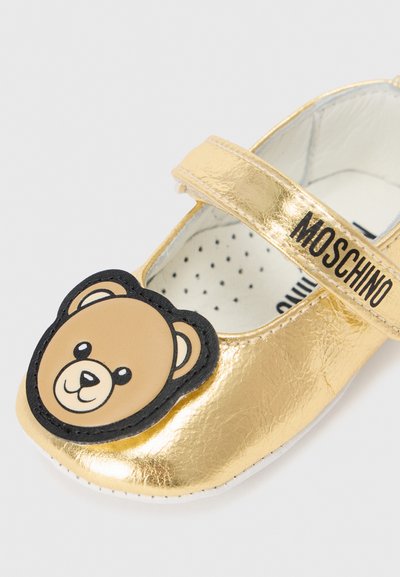 Zapato Mary Jane metálico dorado con adorno de cara de oso negro en la punta. Cuenta con una correa de velcro y una plantilla acolchada con lunares.