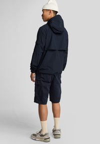 Marineblauwe lichtgewicht jas met capuchon, voorzien van een rugflap, gecombineerd met marineblauwe cargoshorts. Model draagt beige sokken en grijze sneakers.