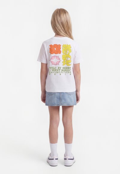 Kind mit langem blondem Haar, das ein weißes T-Shirt mit Grafik und einen hellen Denimrock trägt, steht mit dem Rücken zur Kamera vor weißem Hintergrund.