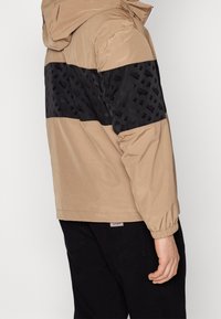 Beżowa kurtka z kapturem z czarną wzorzystą sekcją na klatce piersiowej. Oferuje elastyczne mankiety i prosty dół. Materiał wydaje się gładki.
