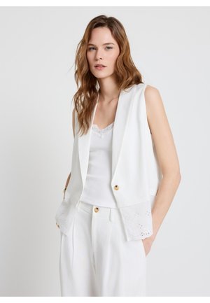 Jeune femme aux cheveux ondulés et lâchés, portant un gilet blanc sans manches avec des boutons sur un débardeur blanc bordé de dentelle et un pantalon blanc.