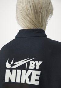 Černá mikina s vysokým límcem, na zadní straně s velkým bílým logem Nike a textem "I BY NIKE". Hladká textura.