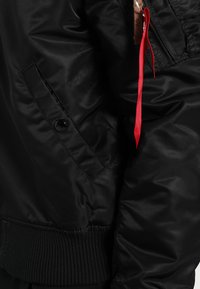 Veste bomber noire avec une texture lisse et brillante, des poignets en tricot et une poche latérale. Dispose d'une tirette de fermeture éclair rouge bien visible et d'accents assortis.