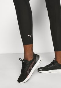 Schwarze Sportleggings mit einem pinken Puma-Logo und schwarzen Sneakers mit weißen Sohlen und pinken Akzenten, die ein schlankes Design zeigen.