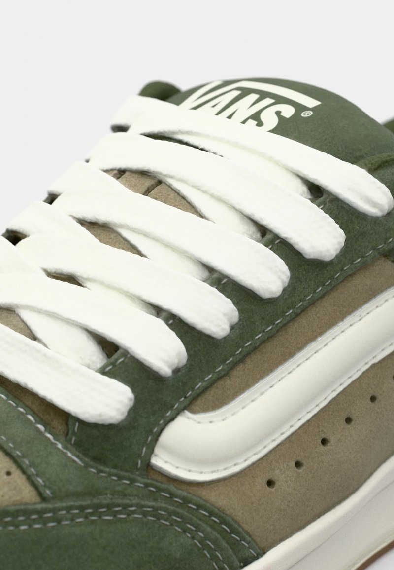 Sneaker in suede verde e beige con lacci bianchi, presenta cuciture in contrasto e una striscia bianca di lato. Logo visibile sulla linguetta.