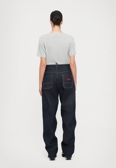 Siva majica s kratkimi rokavi in temno modri široki denim hlače z dvema zadnjima žepoma, ki vključujeta rdečo vezenino na enem žepu.