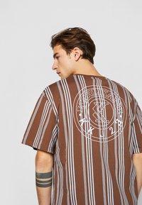 Brun og stripe t-skjorte med blå vertikale linjer, med en sirkulær hvit logo på baksiden. Løs passform med korte ermer og rund hals.