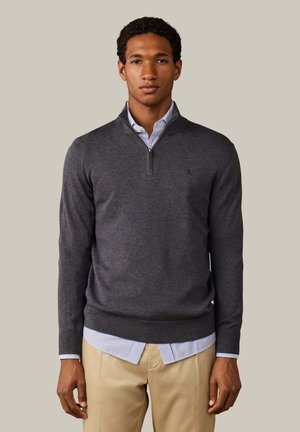 Jonge man draagt een donkergrijze trui met een kwartritssluiting over een blauw gestreept overhemd en beige broek, staand tegen een effen achtergrond.