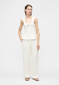 Γυναίκα που φοράει λευκό αμάνικο τοπ και παντελόνες φαρδιά γραμμή με καφέ σανδάλια, στέκεται με το ένα χέρι στην τσέπη μπροστά σε απλό φόντο.