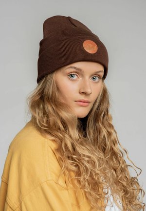 Brązowa dzianinowa czapka z okrągłym pomarańczowym logo, noszona przez osobę z długimi, falującymi blond włosami, wyróżniająca się teksturowanym wzorem materiału.