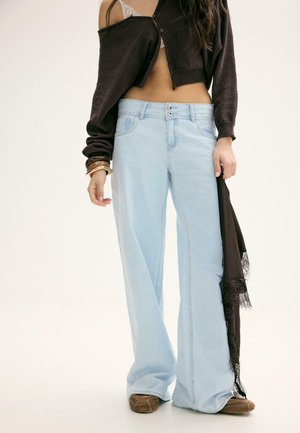 Donna che indossa un cardigan nero corto sopra un reggiseno bianco in pizzo, jeans azzurri a gamba larga, scarpe marroni, che tiene in mano una sciarpa nera rifinita in pizzo.