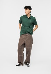 Jonge man staat met de handen achter de rug, draagt een groen poloshirt, losse bruine broek met verstevigde knieën en zwarte sneakers.