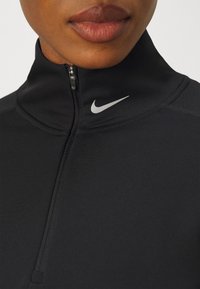 Crna atletska majica s visokim ovratnikom, četvrtim patentnim zatvaračem i srebrnim Nike swoosh logotipom s lijeve strane. Glatka tkanina s prilagođenim dizajnom.