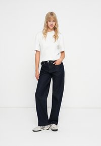 Tamsiai mėlynos plataus kirpimo džinsai, pagaminti iš džinsinio audinio, derinami su balta trumpomis rankovėmis marškenė ir pilkais sportbačiais, stovintys prieš neutralų foną.