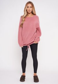 Flauschiger rosa Pullover mit übergroßen Ärmeln und Kordelzugsaum, kombiniert mit schwarzen Leggings und braunen Hausschuhen. Einfaches, lässiges Design.