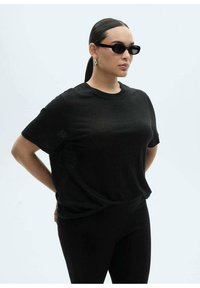 Czarna koszulka na krótki rękaw w luźnym kroju, wykonana z lekkiego materiału, zestawiona z czarnymi legginsami i minimalistycznymi okularami przeciwsłonecznymi.