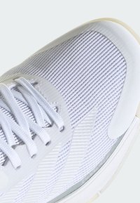 Białe buty sportowe z teksturowaną siateczką, płaskimi sznurowadłami oraz gumową podeszwą z przezroczystą sekcją. Akcenty obejmują szare naszywki.