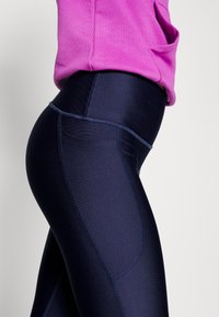 Mørkeblå leggings laget av elastisk stoff med høy livlinje og sidelomme. Har en jevn tekstur og kontrasterende sømmer.